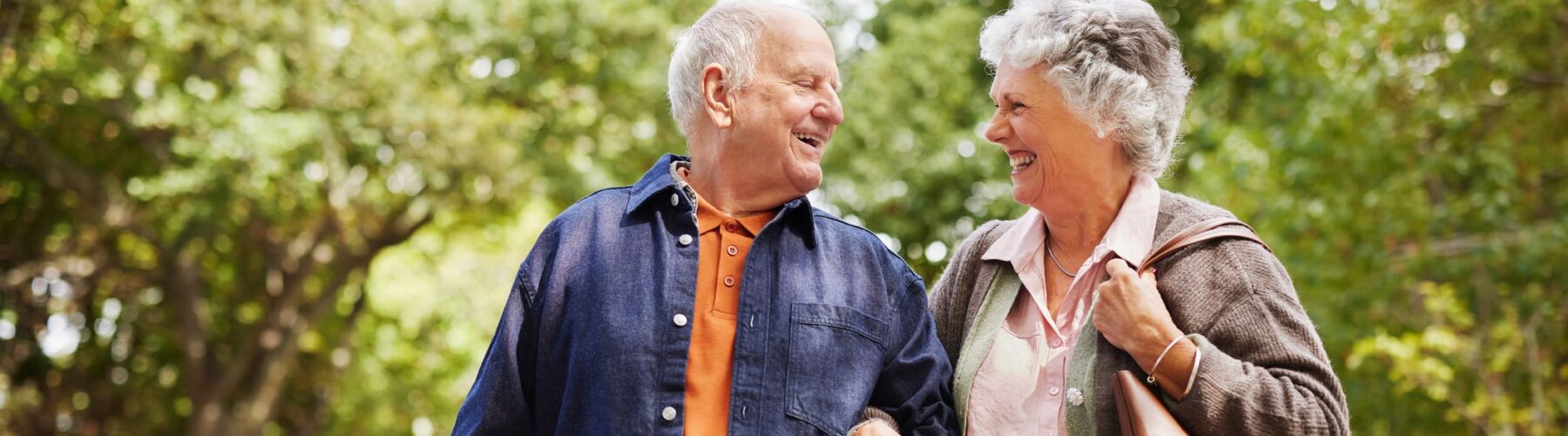 A joyful senior couple walks arm in arm down a tree-lined street, sharing smiles and a warm connection.