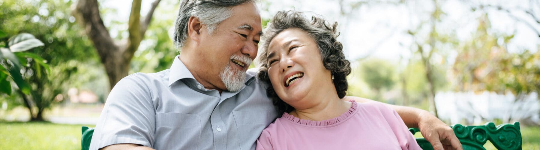 A joyful senior couple sits on a park bench, laughing and sharing a loving moment surrounded by lush greenery.
