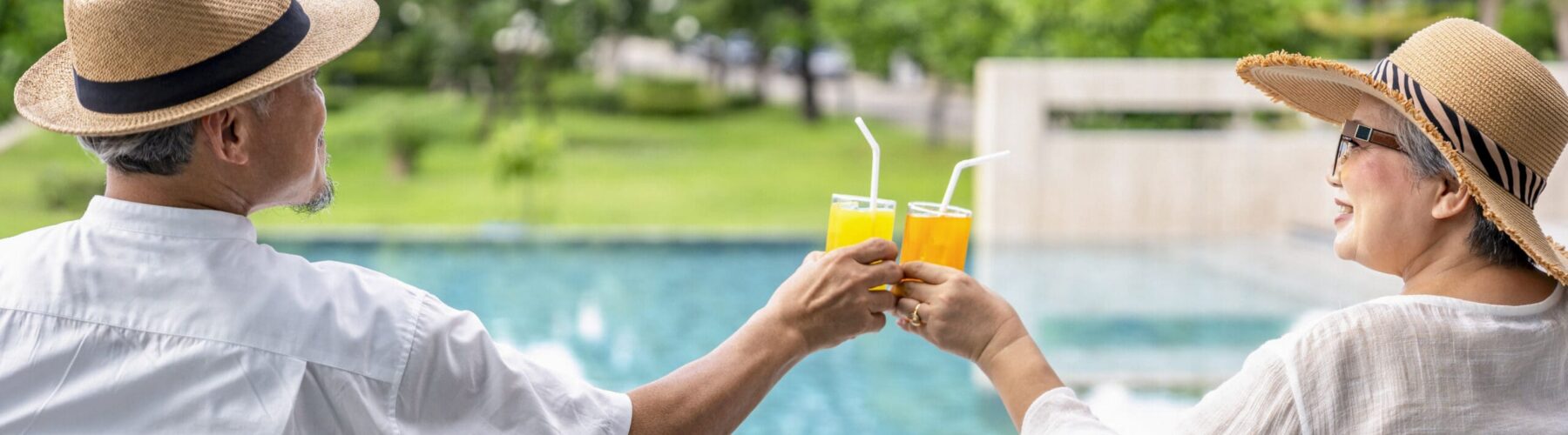 Two seniors enjoy a toast with orange drinks by a sparkling pool, surrounded by lush greenery on a sunny day.