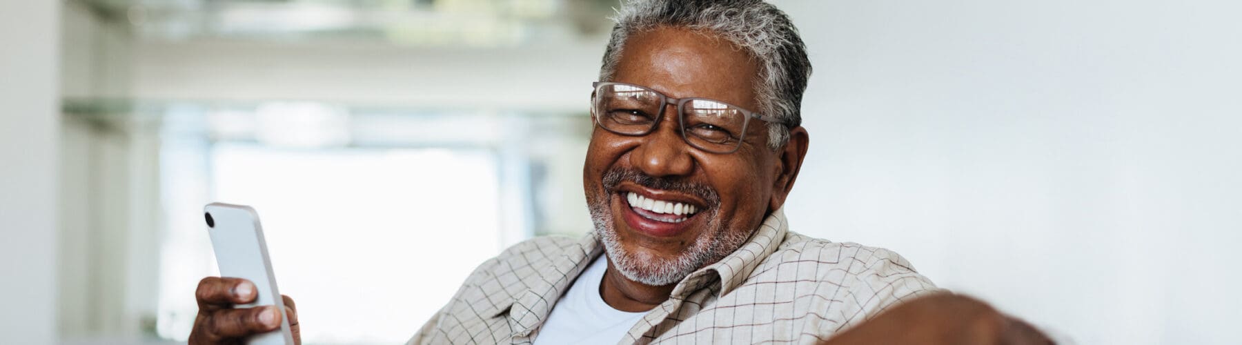 Joyful senior man with glasses smiles while holding a smartphone in a stylish, bright living space.