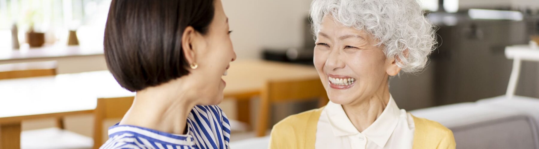 Joyful interaction between a woman in a striped shirt and an older woman in a yellow cardigan, sharing laughter in a cozy setting.
