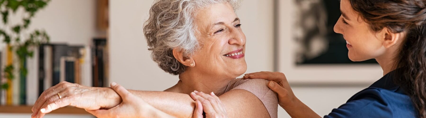A caregiver assists a senior woman with gentle stretching in a bright, welcoming room filled with greenery and books.