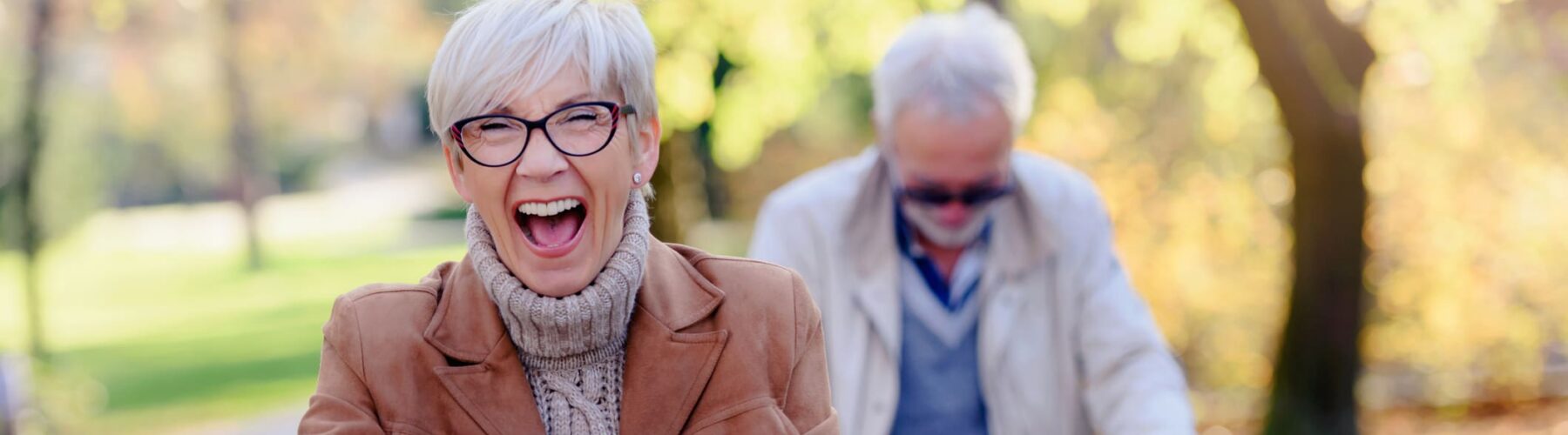 Cheerful active senior couple with bicycle in public park together having fun - Grace Management Joyful senior woman riding a bike in a sunny park, while a man in the background enjoys the vibrant autumn scenery.