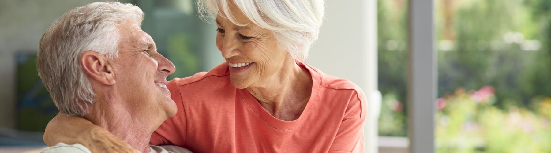 Head And Shoulders Of Loving Senior Couple On Hugging Indoors At Home Together - Grace Management Joyful seniors sharing a tender moment, surrounded by lush greenery in a bright, inviting living space.
