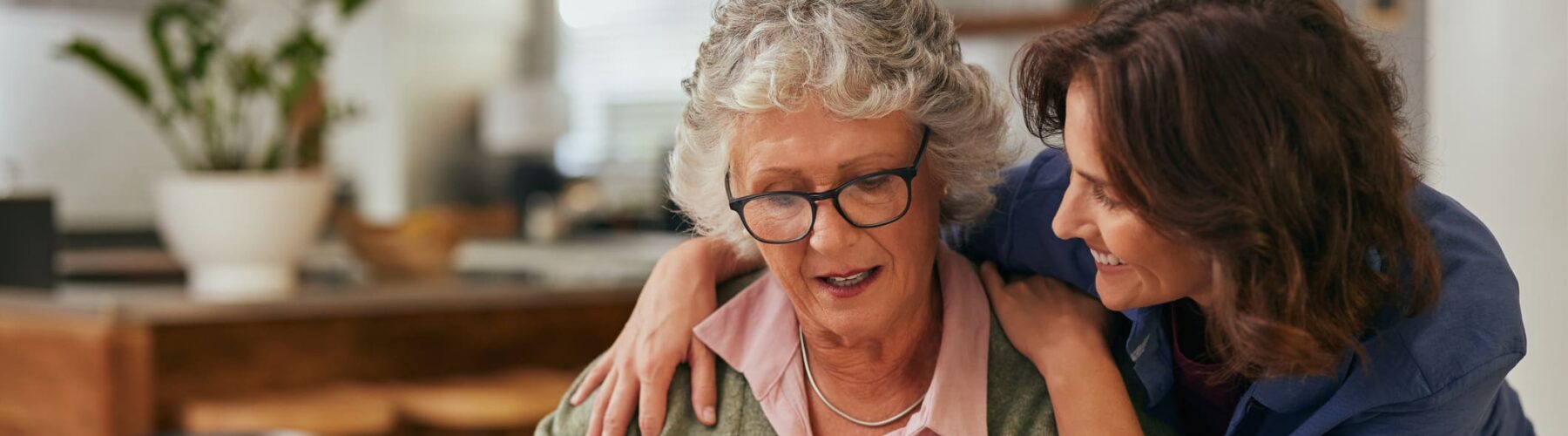A smiling woman assists a senior resident with a puzzle, embodying warmth and supportive companionship in a cozy kitchen setting.