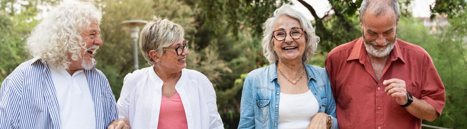 Four smiling seniors stroll together in a lush park, sharing joyful moments in a vibrant community setting.