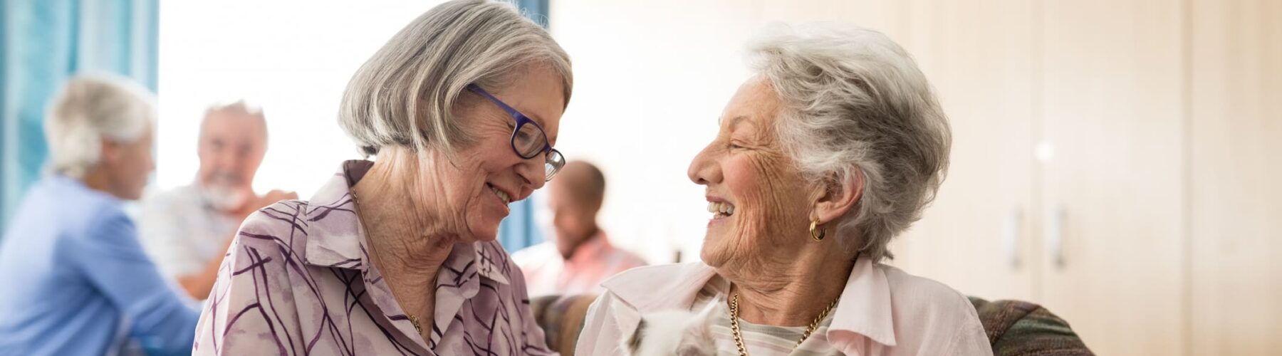 Two smiling senior women share a joyful moment while holding a small pet in a cozy, engaging communal space.