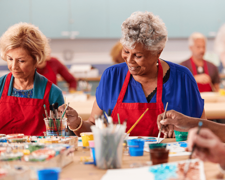 Two smiling older women in red aprons paint at a craft table with brushes and colored paints in a bright classroom setting.