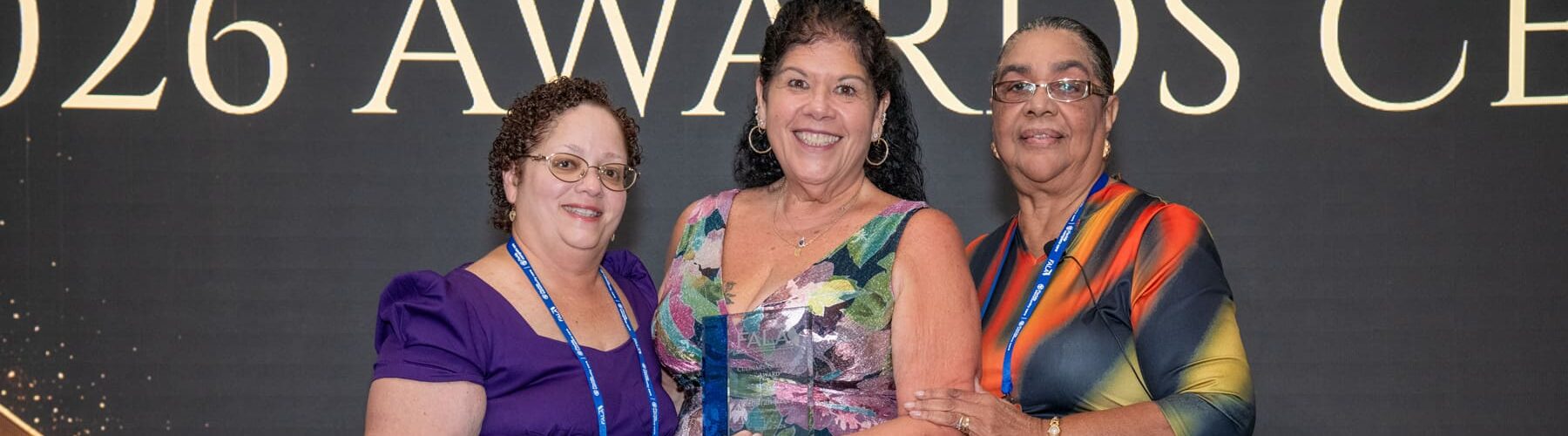 Three women proudly pose together, celebrating an award presentation, with a backdrop displaying 2026 AWARDS CEREMONY.