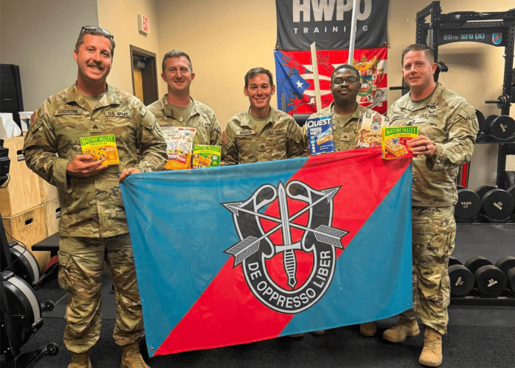 Five military personnel proudly hold snacks in front of a flag, showcasing camaraderie and strength in a fitness environment.