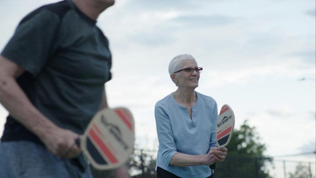 Two seniors stand outside with paddles playing pickle ball - Grace Management Two seniors stand outside with paddles, playing pickle ball.