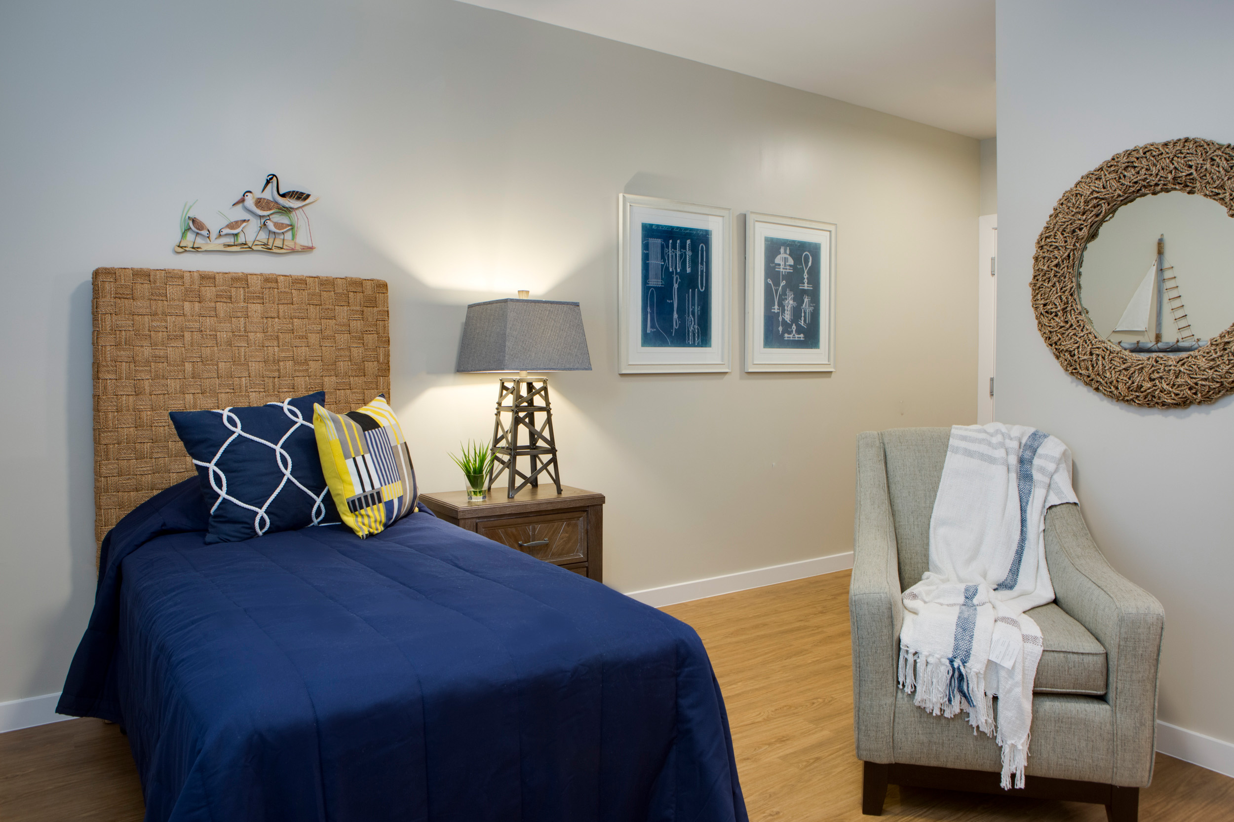 Heritage-Oaks1826-1 - Grace Management A bedroom with a twin-sized bed with blue bedding and throw pillows, a bedside table with a lamp, and a gray sofa chair with a white throw blanket on it.
