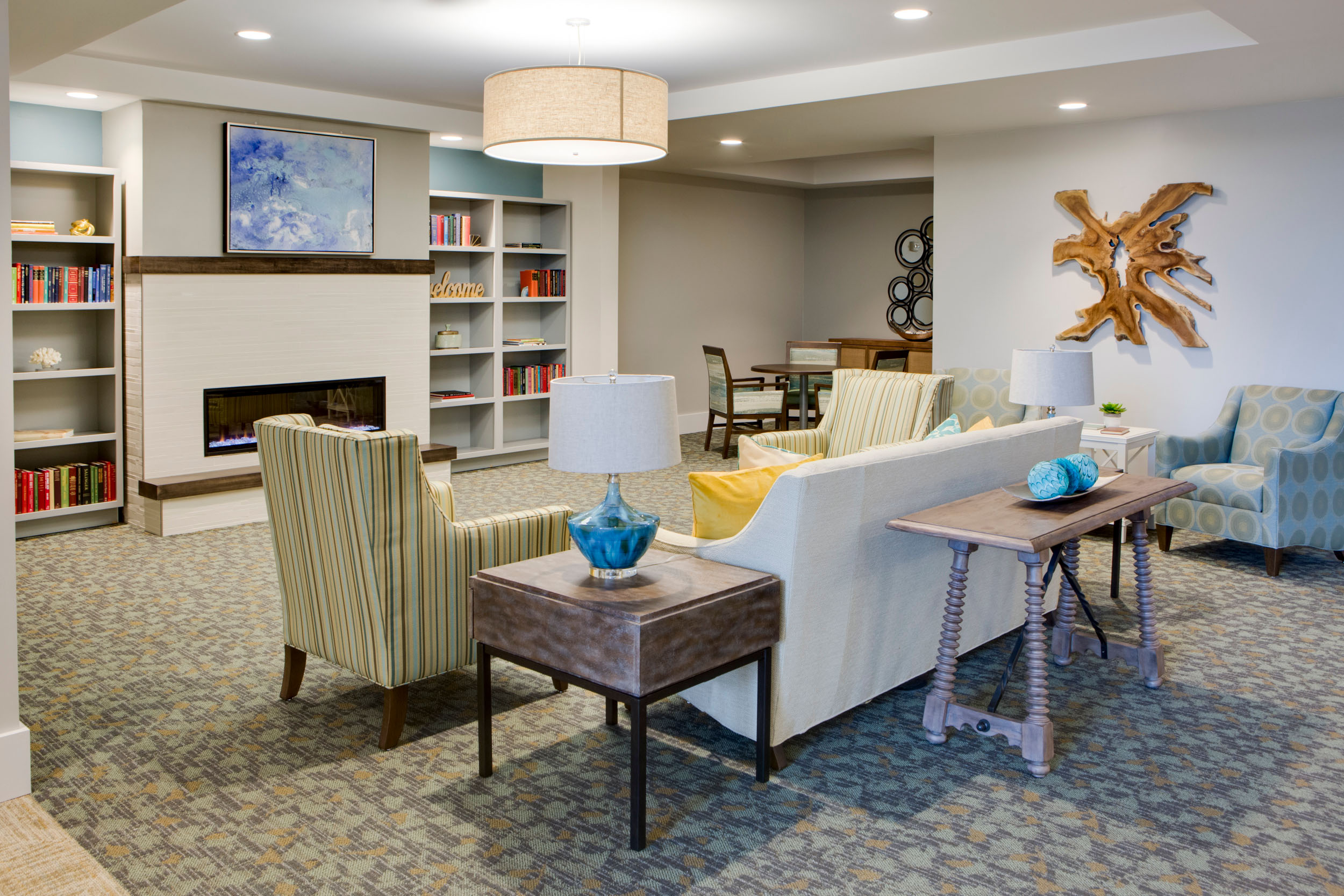 Heritage-Oaks1836 - Grace Management A living room are with a white couch and throw pillows on it, sofa chairs next to it with side tables and lamps on it, located across a digital fireplace that's underneath a wall painting, which is next to two bookshelves with books on them at Heritage Oaks of Englewood, a senior living community near Venice, FL.