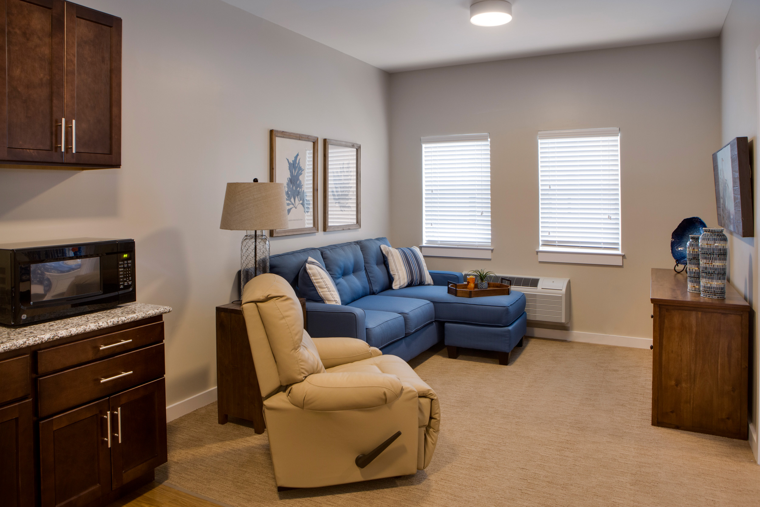 Heritage-Oaks1848 - Grace Management A living room with a blue couch facing the TV that's mounted to the wall underneath a dresses, with a mustard-colored reclining sofa chair next to the couch.