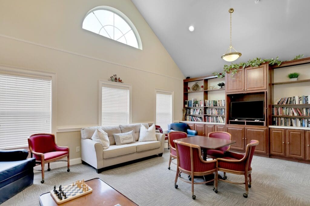 MG_7957-1024x683 - Grace Management An indoor sitting area with white couch, a blue sofa chair, a brown table with pink cushioned chairs around it, all located in front of a long bookshelf full of books and a TV.