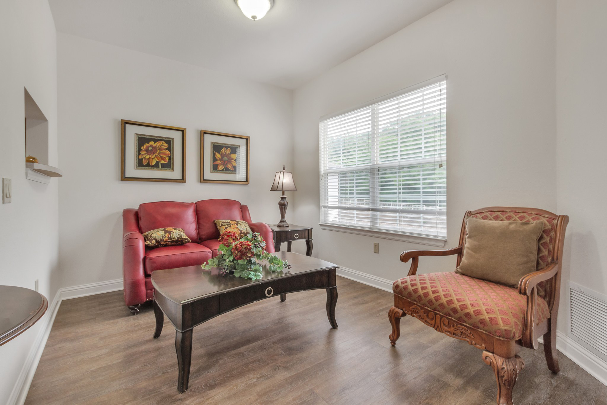 A living room area with a red leather couch and flower-patterned throw pillows on it, a side table with a lamp, a coffee table with a flower bouquet on it, and a sofa chair next to it.