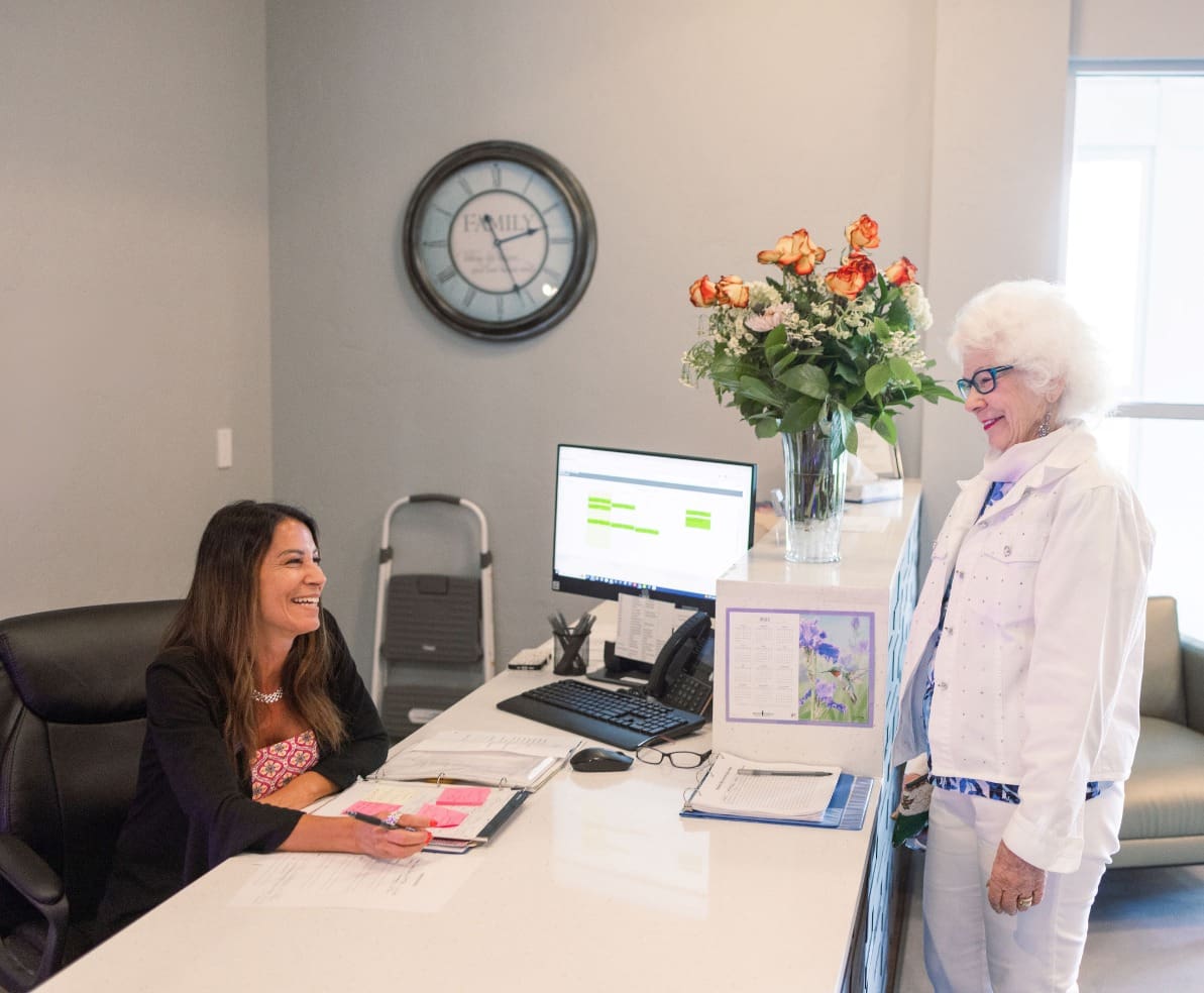 1 - Grace Management Senior woman stands at the reception desk, chatting with a staff member on the other side at The Summit of Coralville.