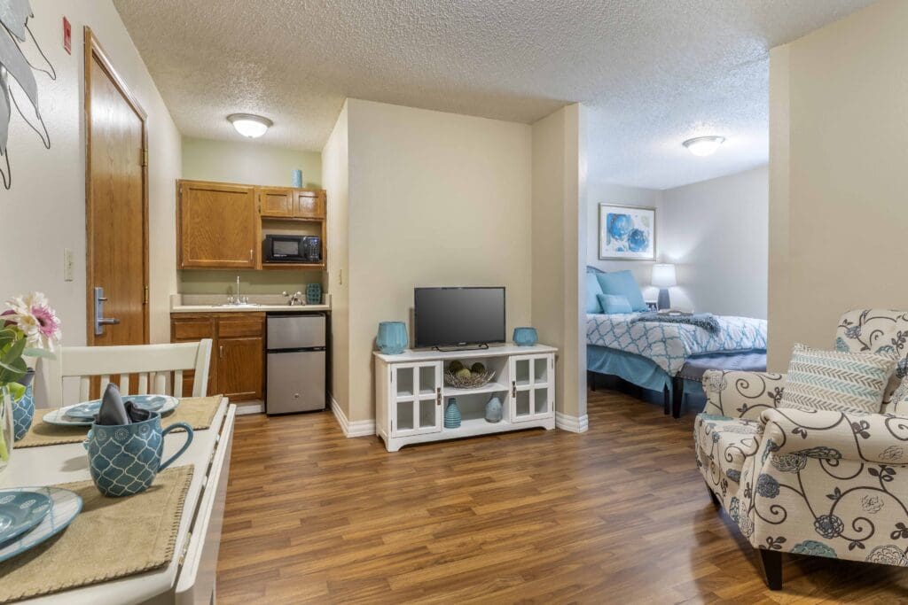 Final-Selects-cs3-8-1024x683 - Grace Management Interior image of a resident's room with a small kitchenette, living room area, and a bed in the background at Inspirations of Bartlesville.