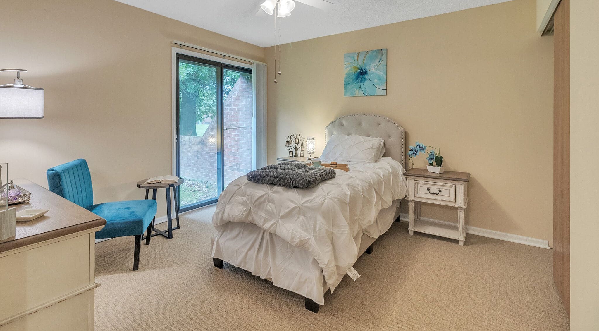 IMG_0019-e1687292737673 - Grace Management A bedroom with a twin-sized bed with white bedding and a gray throw blanket, a painting of a blue flower above the bed, and a blue chair next to it.